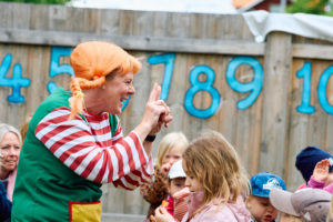 En förskolelärare utklädd till Pippilångstrum står ute på förskolans gård med glada barn omkring sig och sjunger sånger med gester.
