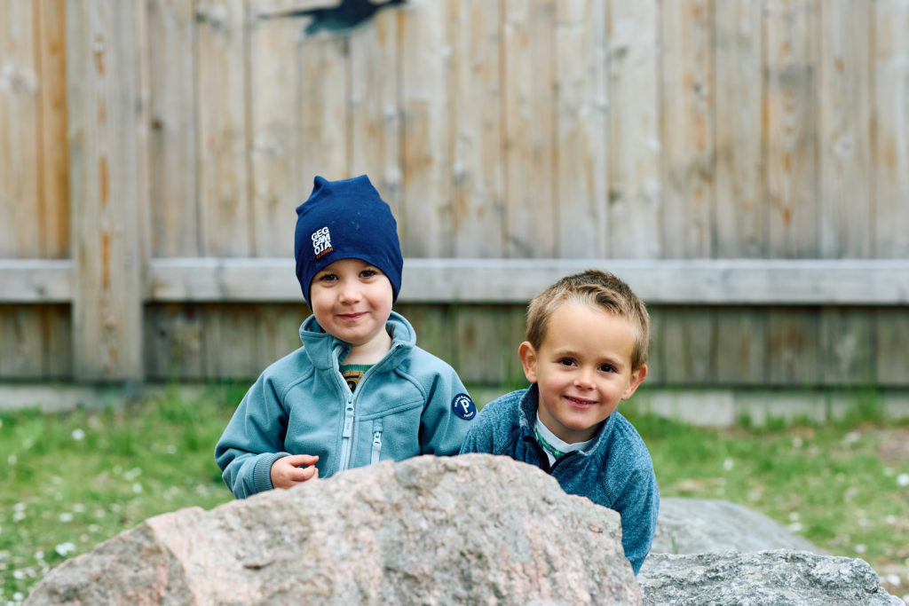 Två pojkar kikar fram bakom en sten och tittar busigt in i kameran.